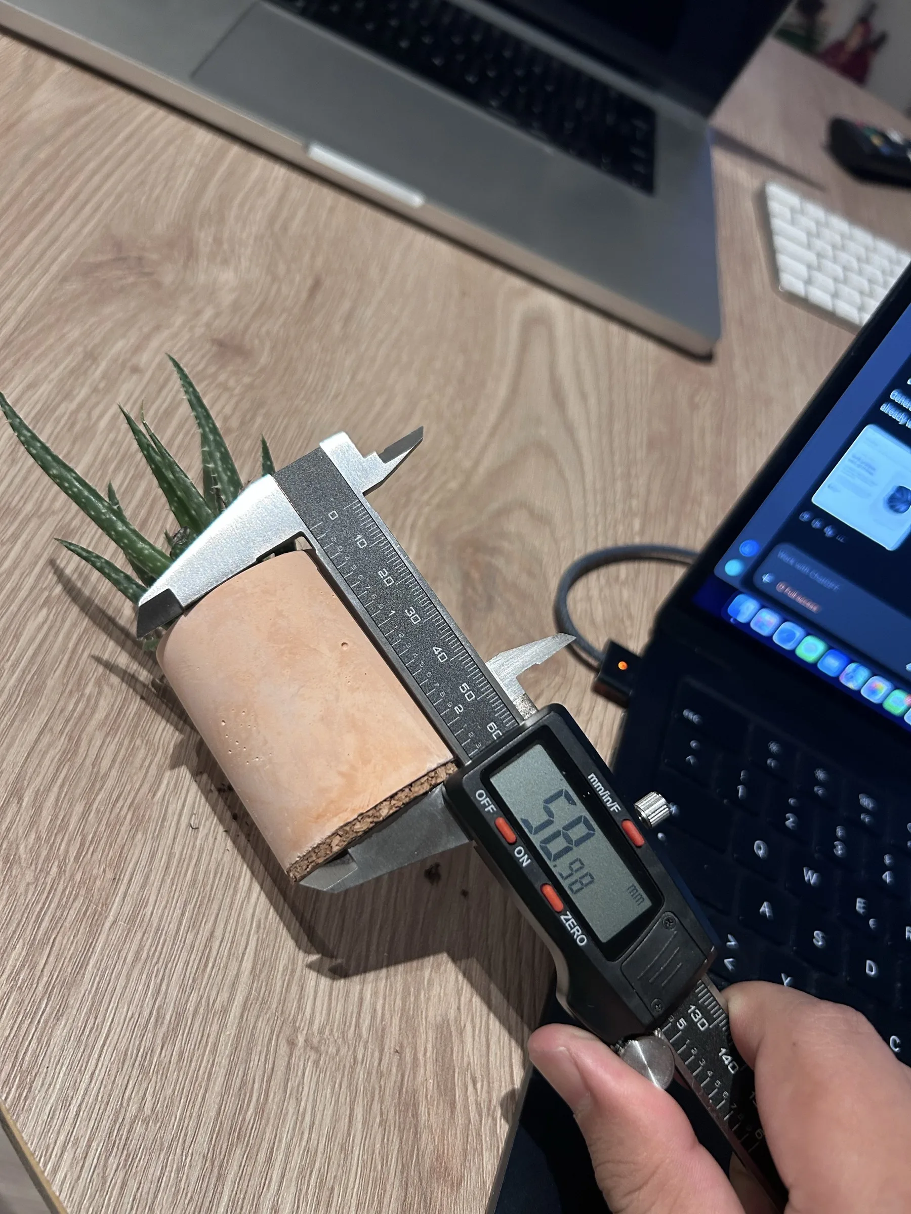 The caliper measuring the peach-coloured pot and cork base vertically, with its digital display reading 58.98 millimetres.